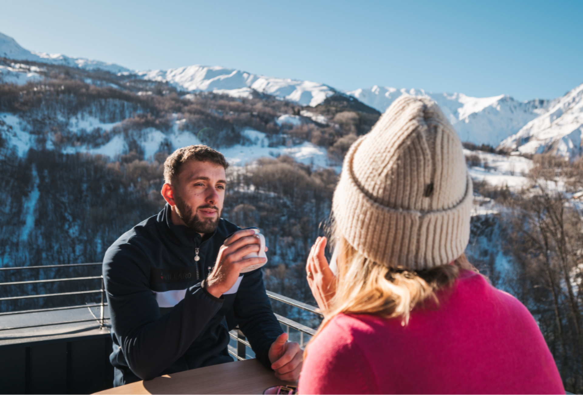 Un couple souriant profite d’un moment en terrasse dans une station de ski, entouré de montagnes enneigées.
